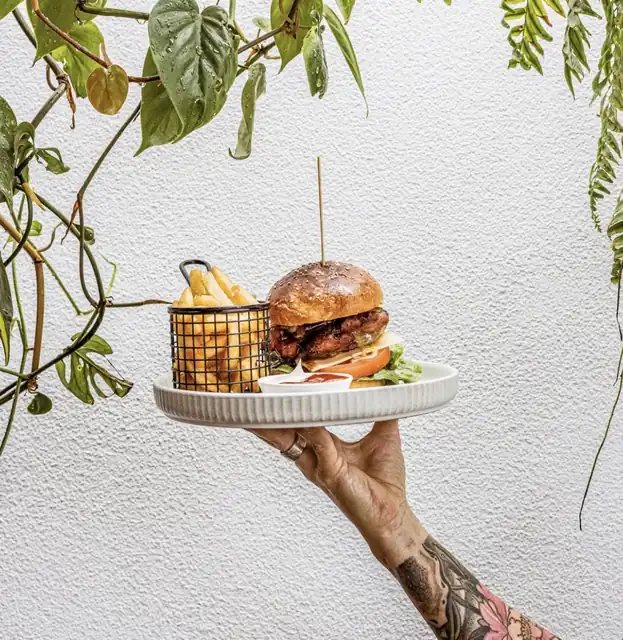 Beef and bacon burger with a basket of chips on a plate held high against a white wall