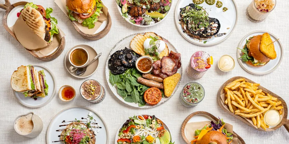 Overhead spread of brunch plates, salad bowls, a burger and golden chips on a white linen table