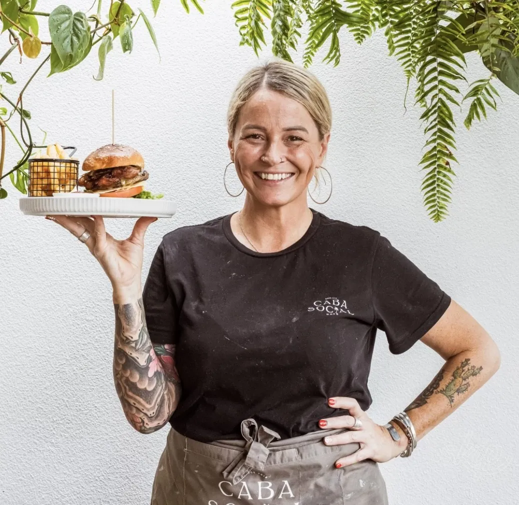 Team member in the cafe tee and apron holding up a plate of burger and chips beneath hanging ferns